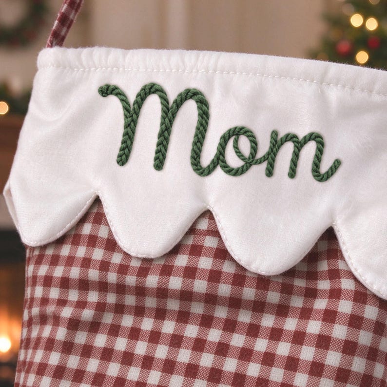 May include: A Christmas stocking featuring a red and white gingham pattern on the main body. The top cuff is white with a scalloped edge and the word Mom embroidered in dark green rope-style lettering across the center of the cuff.