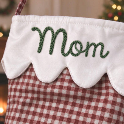 May include: A Christmas stocking featuring a red and white gingham pattern on the main body. The top cuff is white with a scalloped edge and the word Mom embroidered in dark green rope-style lettering across the center of the cuff.