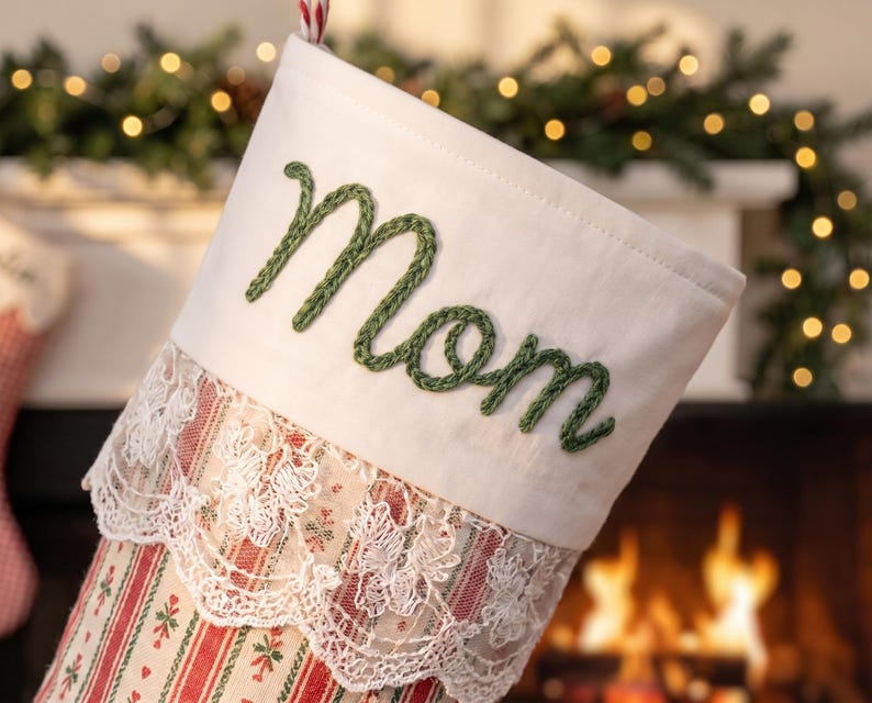 May include: A Christmas stocking featuring a white cuff with the word Mom embroidered in green yarn. Below the cuff is a layer of white lace trim. The body of the stocking has a red and white striped pattern with small green leaf accents.