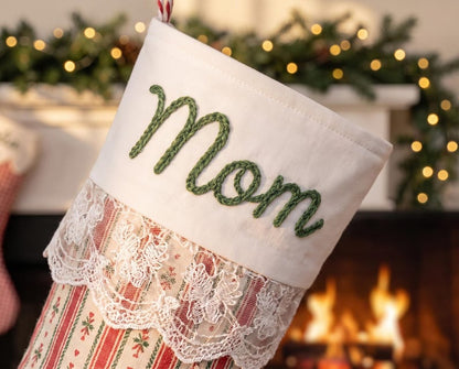 May include: A Christmas stocking featuring a white cuff with the word Mom embroidered in green yarn. Below the cuff is a layer of white lace trim. The body of the stocking has a red and white striped pattern with small green leaf accents.